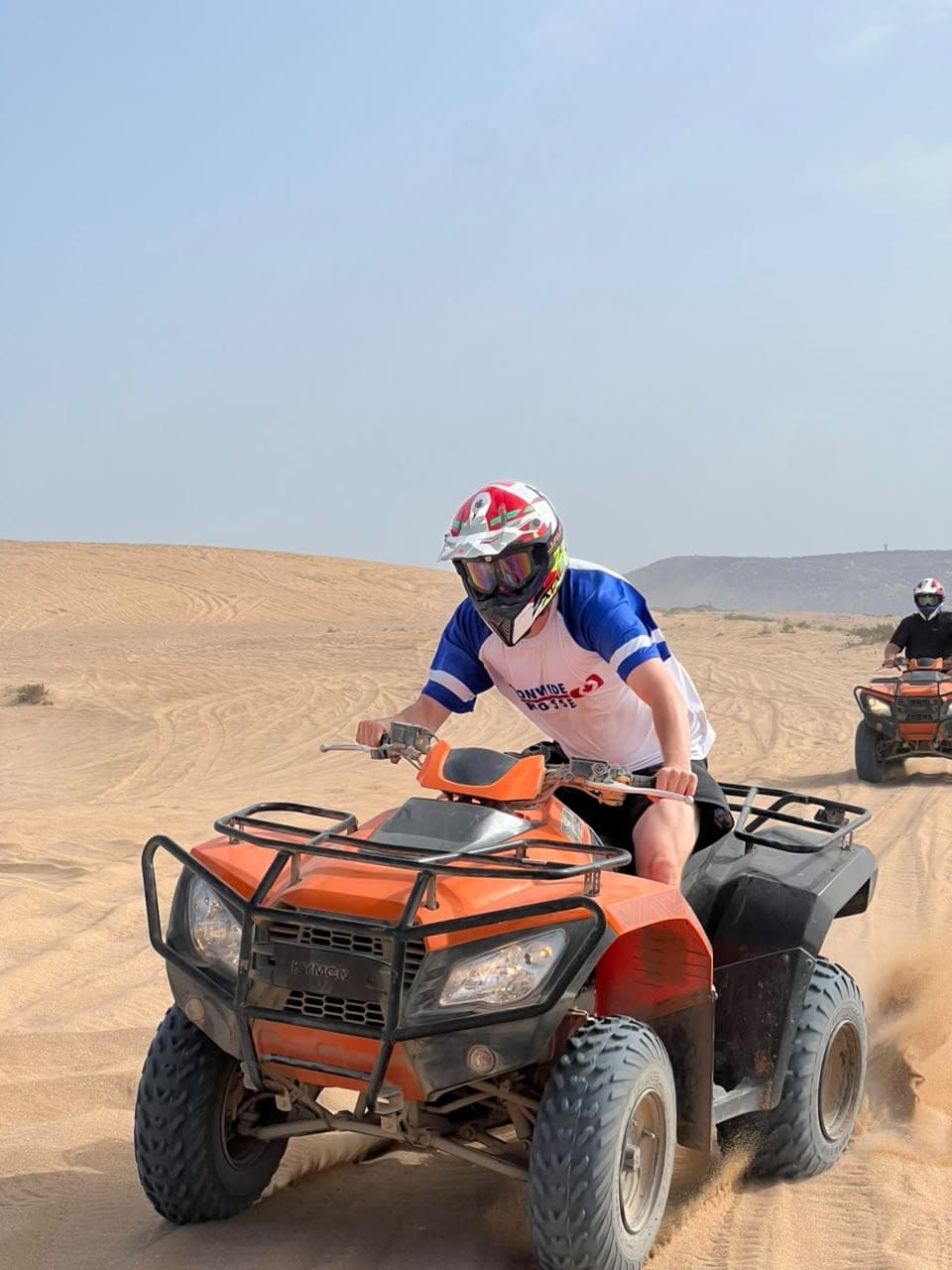 Rider in helmet and goggles driving a quad bike through the sand, kicking up dust, in the Timlalin dunes